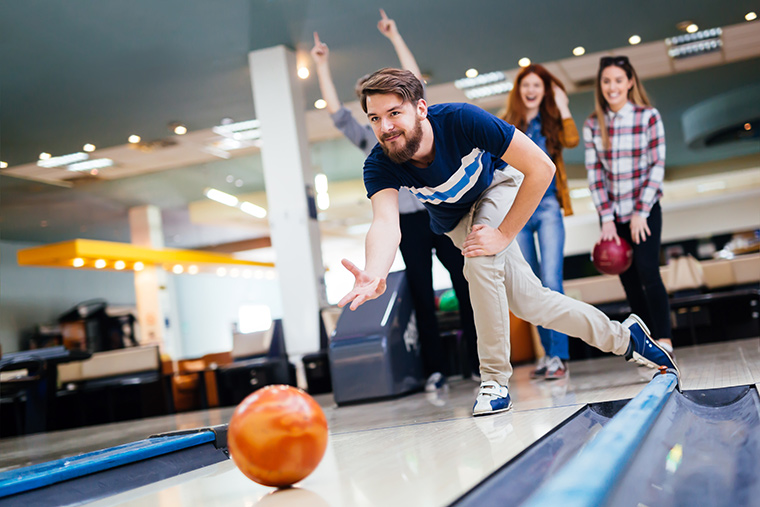 Man launching bowling ball with friends in the background cheering him on.