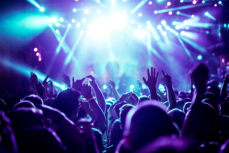 A lively concert scene featuring enthusiastic attendees with hands raised in the air, illuminated by pulsating strobe lights.